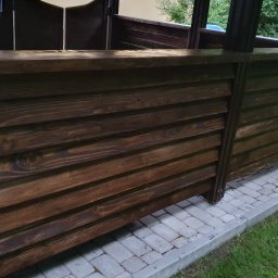 F.H.U. TreeBiel Piotr Bielak - Fragment konstrukcji z ciemnego, impregnowanego drewna, prawdopodobnie elementu altany lub pergoli, widoczny na tle kostki brukowej i trawy.