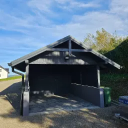 Ciemnoszara, drewniana konstrukcja zadaszenia w ogrodzie, z kostką brukową na podłożu, widoczne gniazdka elektryczne, błękitne niebo w tle.