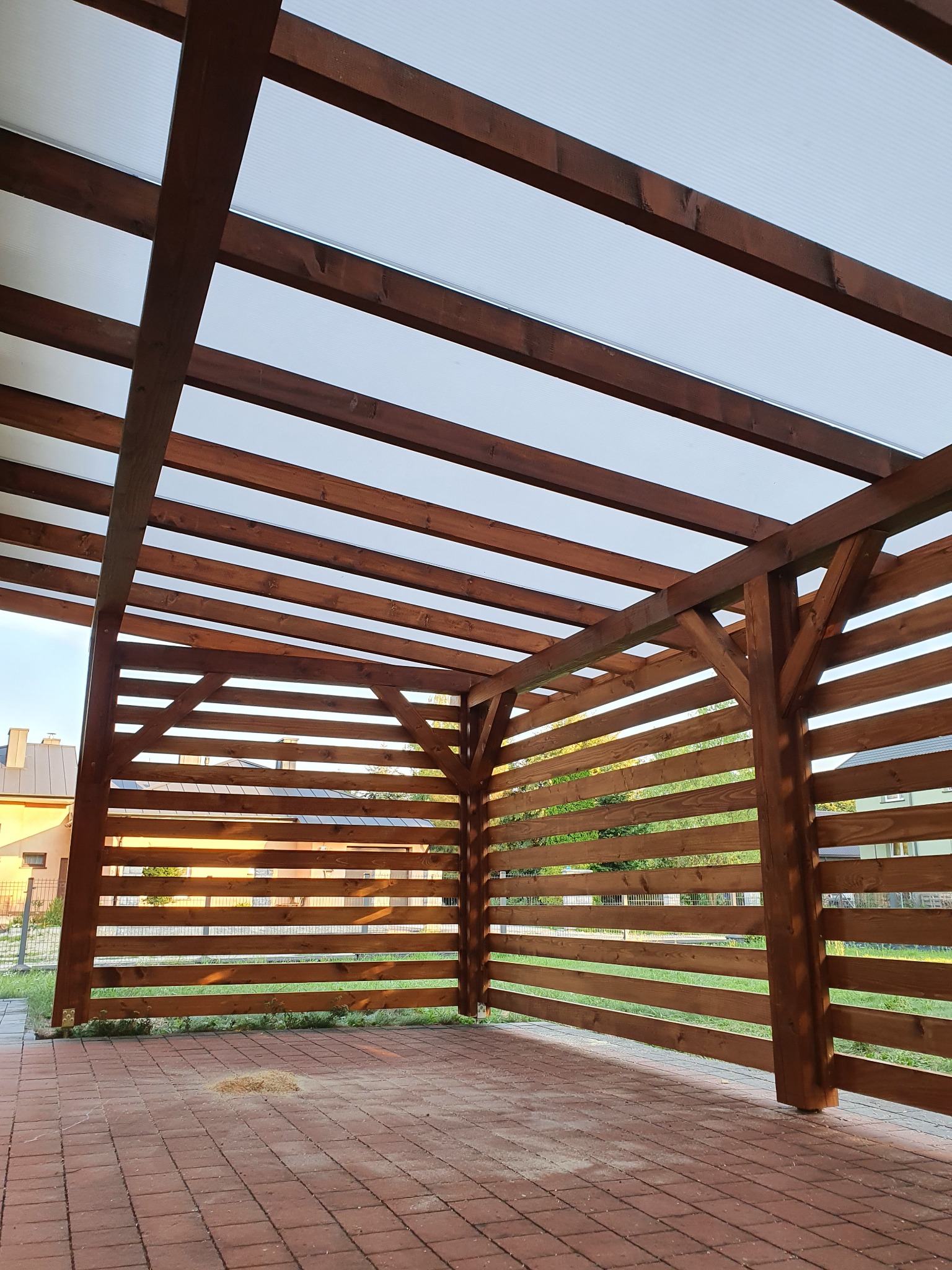 Konstrukcja z ciemnego drewna z zadaszeniem z przezroczystego tworzywa, tworząca częściowo osłoniętą przestrzeń na posesji, widok od wewnątrz, na podłożu widoczne ślady po obróbce drewna.