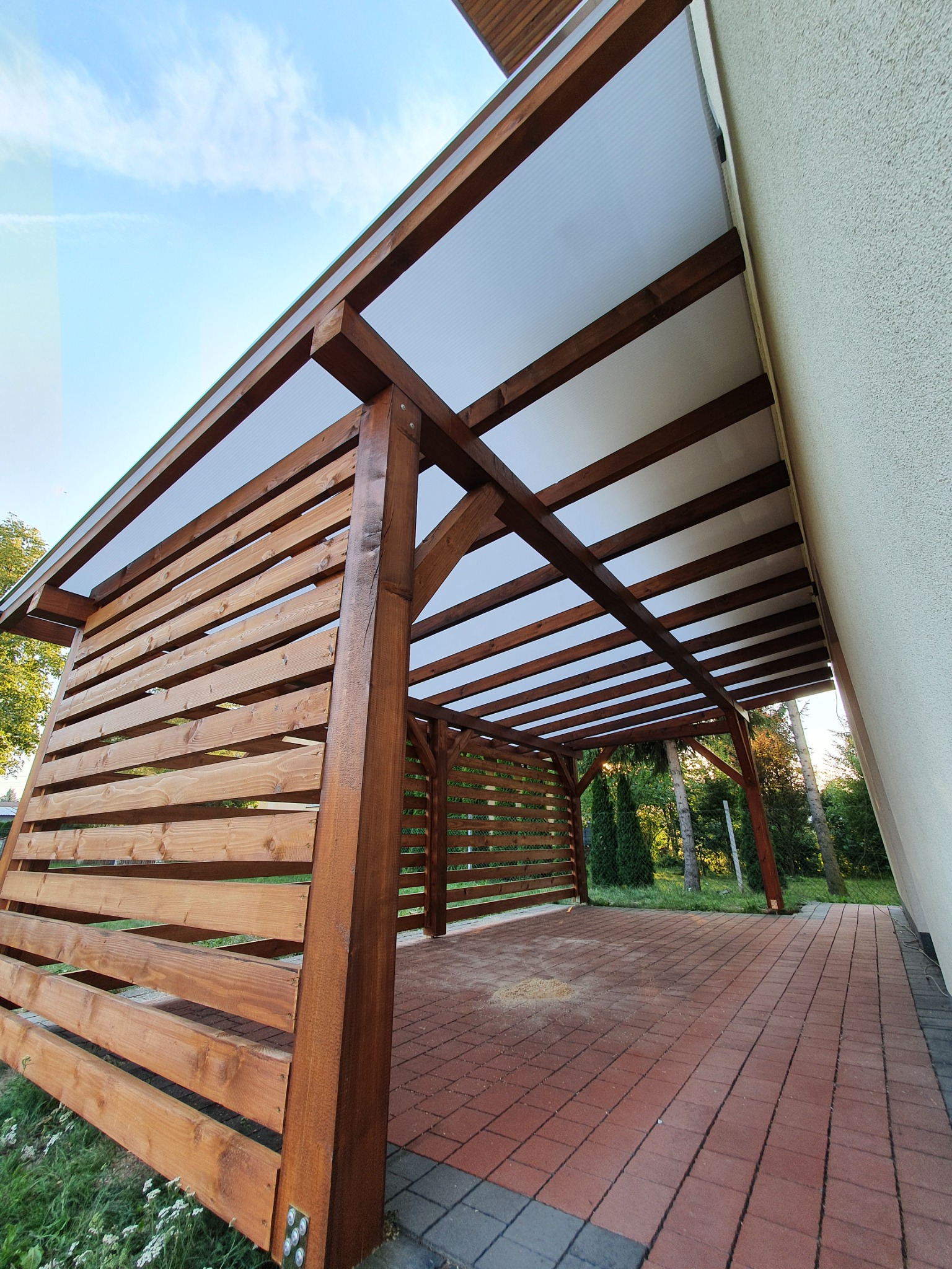 Zadaszona konstrukcja z drewna z ażurowymi ścianami i przezroczystym dachem, przylegająca do budynku z kostką brukową na podłożu.