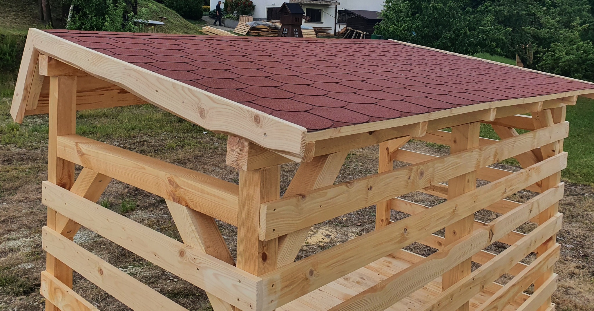 Surowa drewniana konstrukcja z czerwonym dachem z gontu bitumicznego, widoczna na tle trawnika i zabudowań.