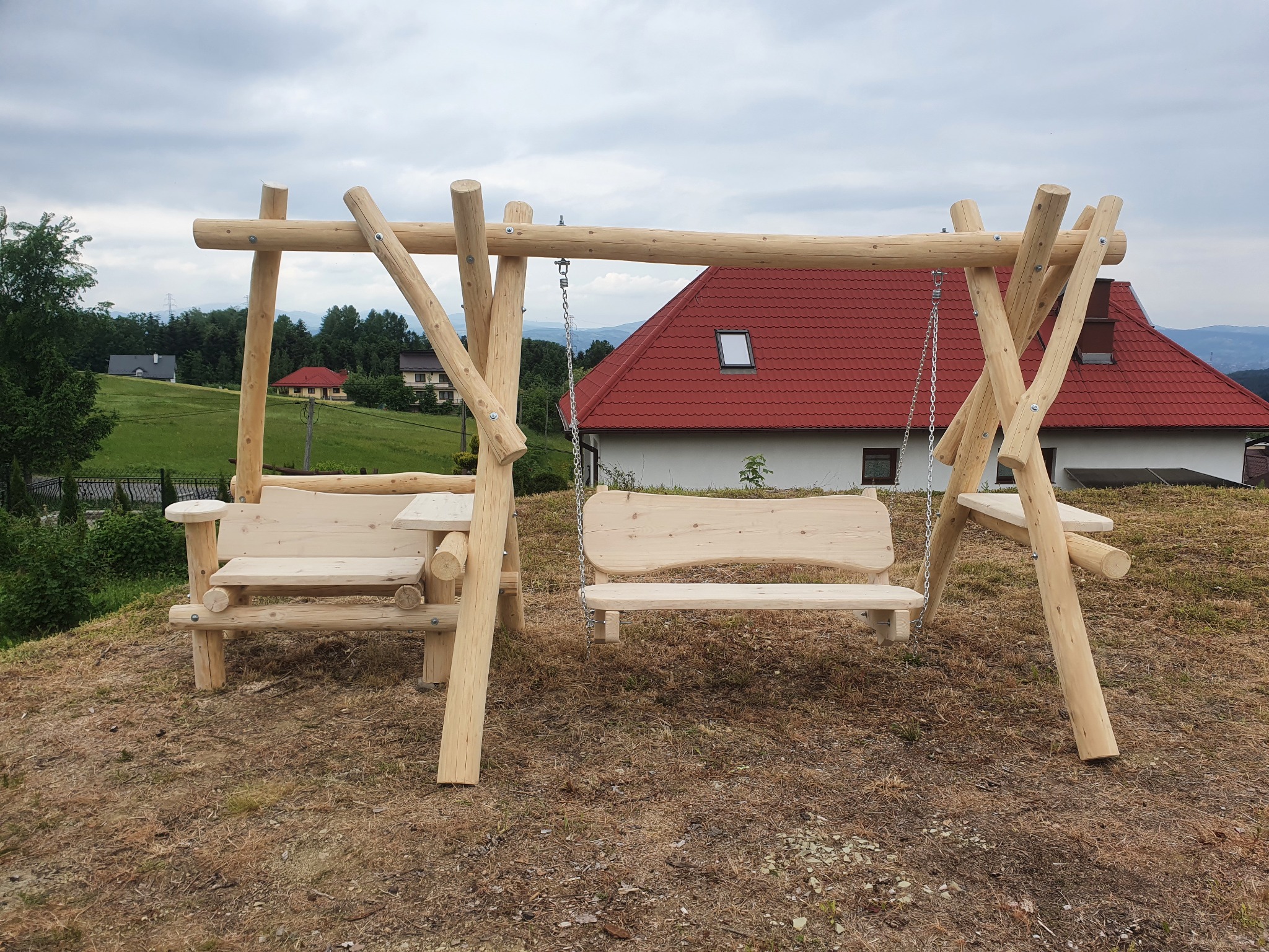 Drewniana huśtawka ogrodowa z dwoma siedziskami i stolikami bocznymi, wykonana z okrąglaków, na tle domu z czerwoną dachówką i pochmurnego nieba.