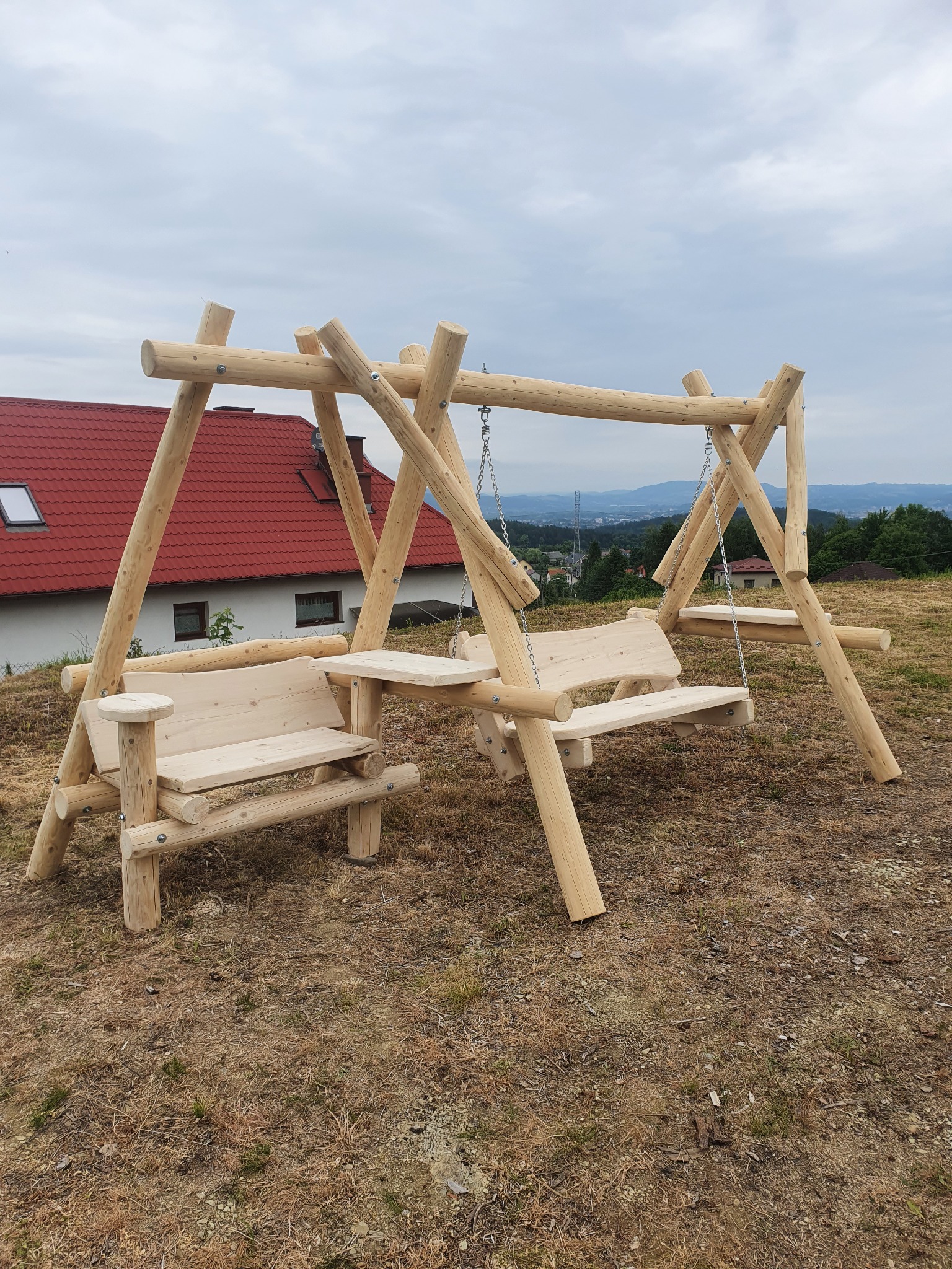 Drewniana huśtawka ogrodowa z dwoma siedziskami i małym stolikiem, wykonana z okrąglaków, na tle domu z czerwoną dachówką i widokiem na panoramę miasta.