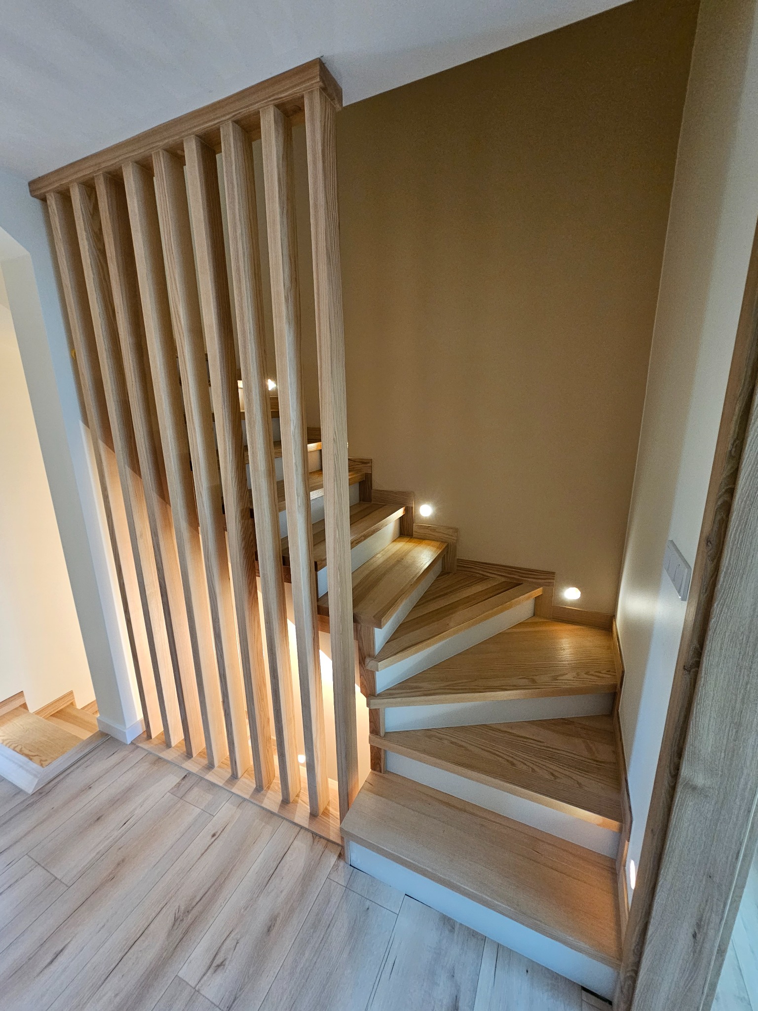 Drewniane schody z oświetleniem LED i ażurową balustradą z pionowych listew, widoczne z góry, na tle beżowej ściany. Nowoczesny design, jasne drewno.