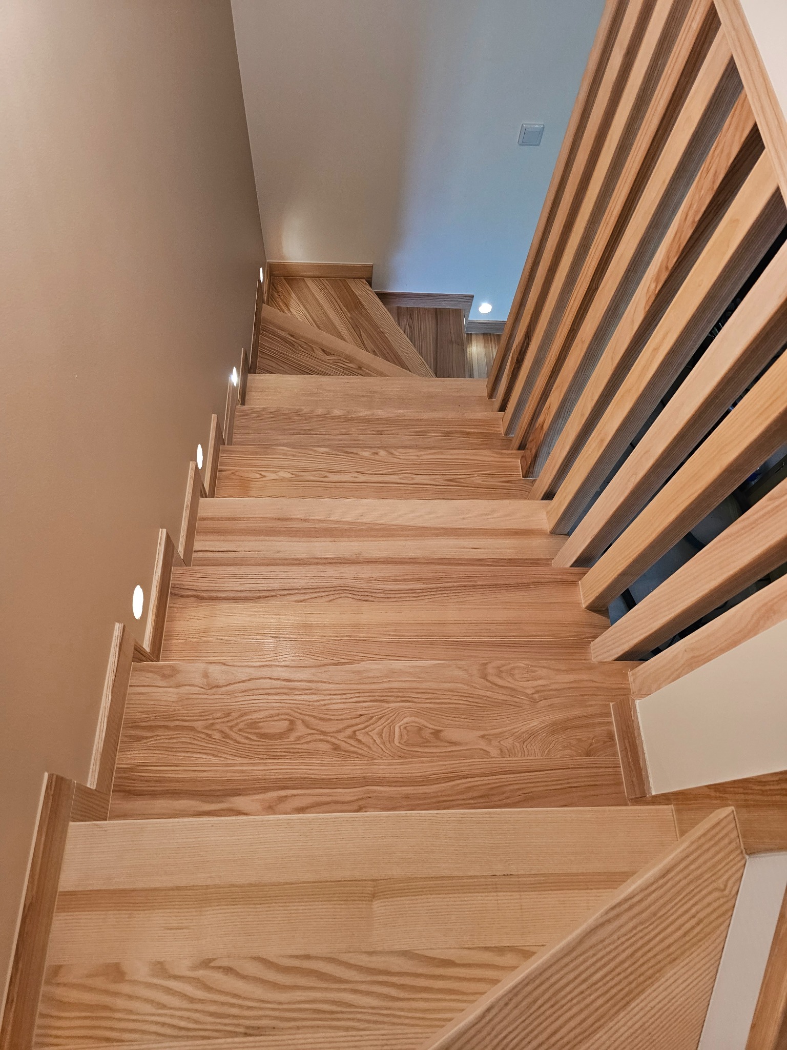 Nowoczesne schody drewniane z oświetleniem punktowym i ażurową balustradą, widok z góry, ukazujący naturalny rysunek drewna i minimalistyczny design.