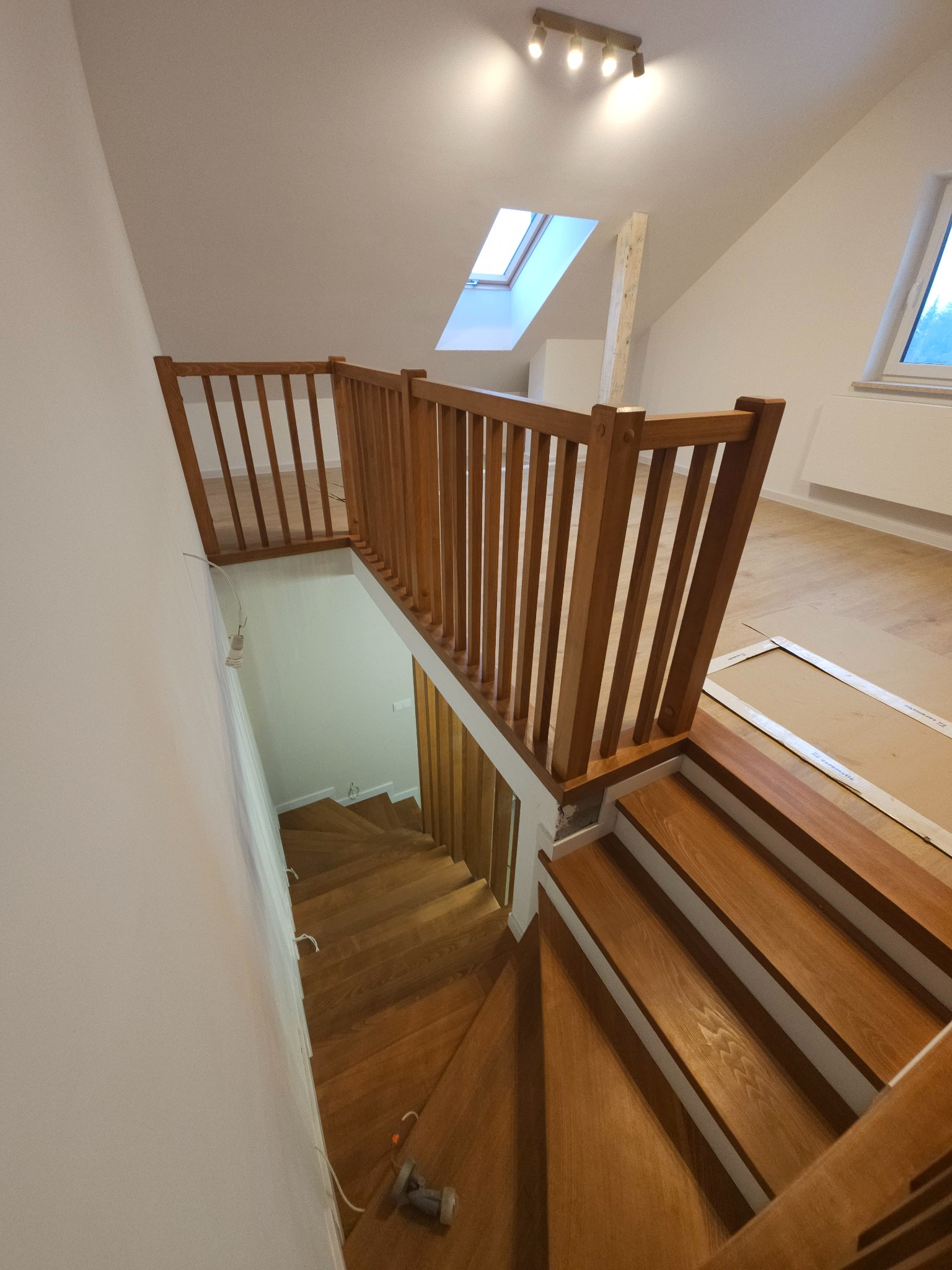 Jasne, drewniane schody z balustradą w domu z poddaszem. Widok z góry, ukazujący detale wykończenia i naturalne oświetlenie z okna dachowego. Minimalistyczny design.