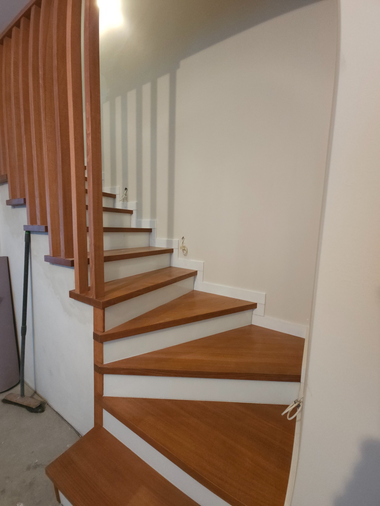 Spiralne, drewniane schody z balustradą w Nowym Sączu. Stopnie w ciepłym odcieniu brązu kontrastują z białymi podstopnicami. Widoczne detale wykończenia i naturalne oświetlenie.