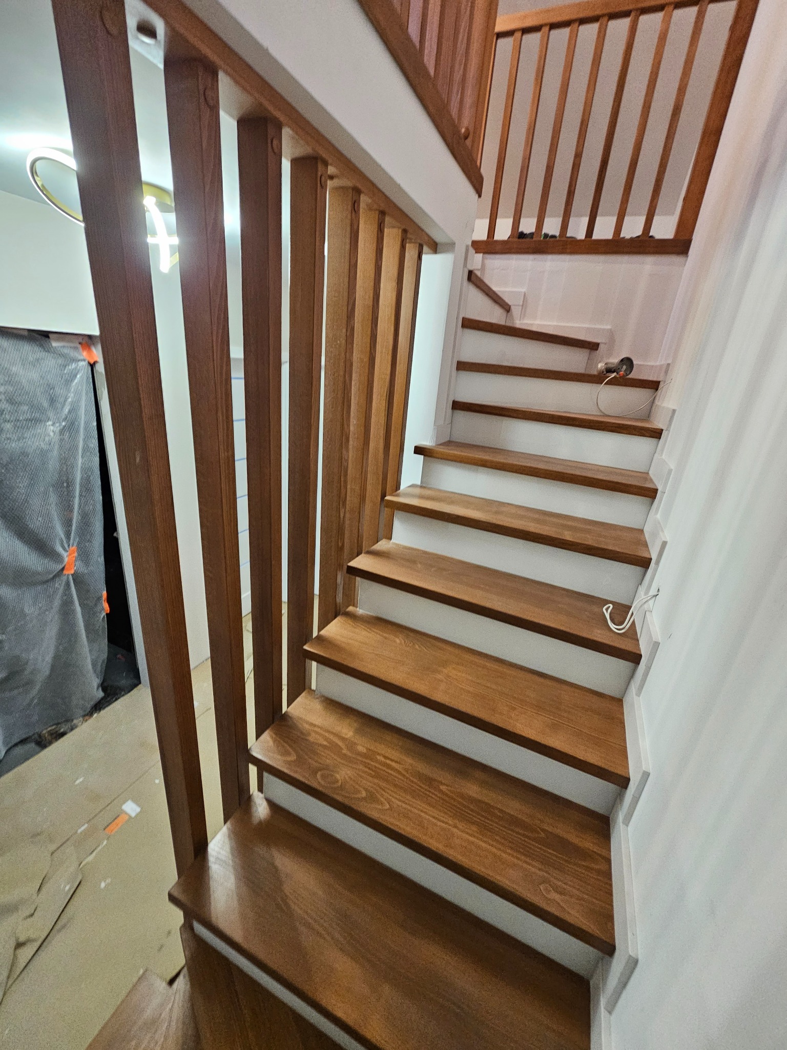 Nowoczesne schody drewniane z balustradą z pionowych listew, wykończone w ciepłym odcieniu brązu, z białymi podstopnicami. Widoczne narzędzia i zabezpieczenia podłogi.