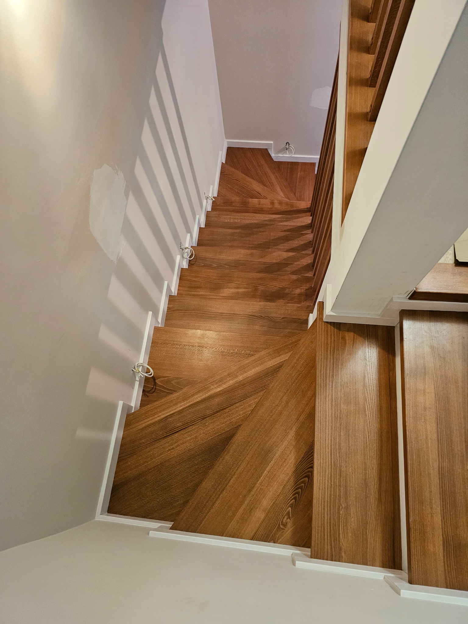 Nowoczesne schody z drewna o unikalnym wzorze ułożenia klepek, widok z góry. Białe ściany i balustrada kontrastują z ciepłym odcieniem drewna. Minimalistyczny design.