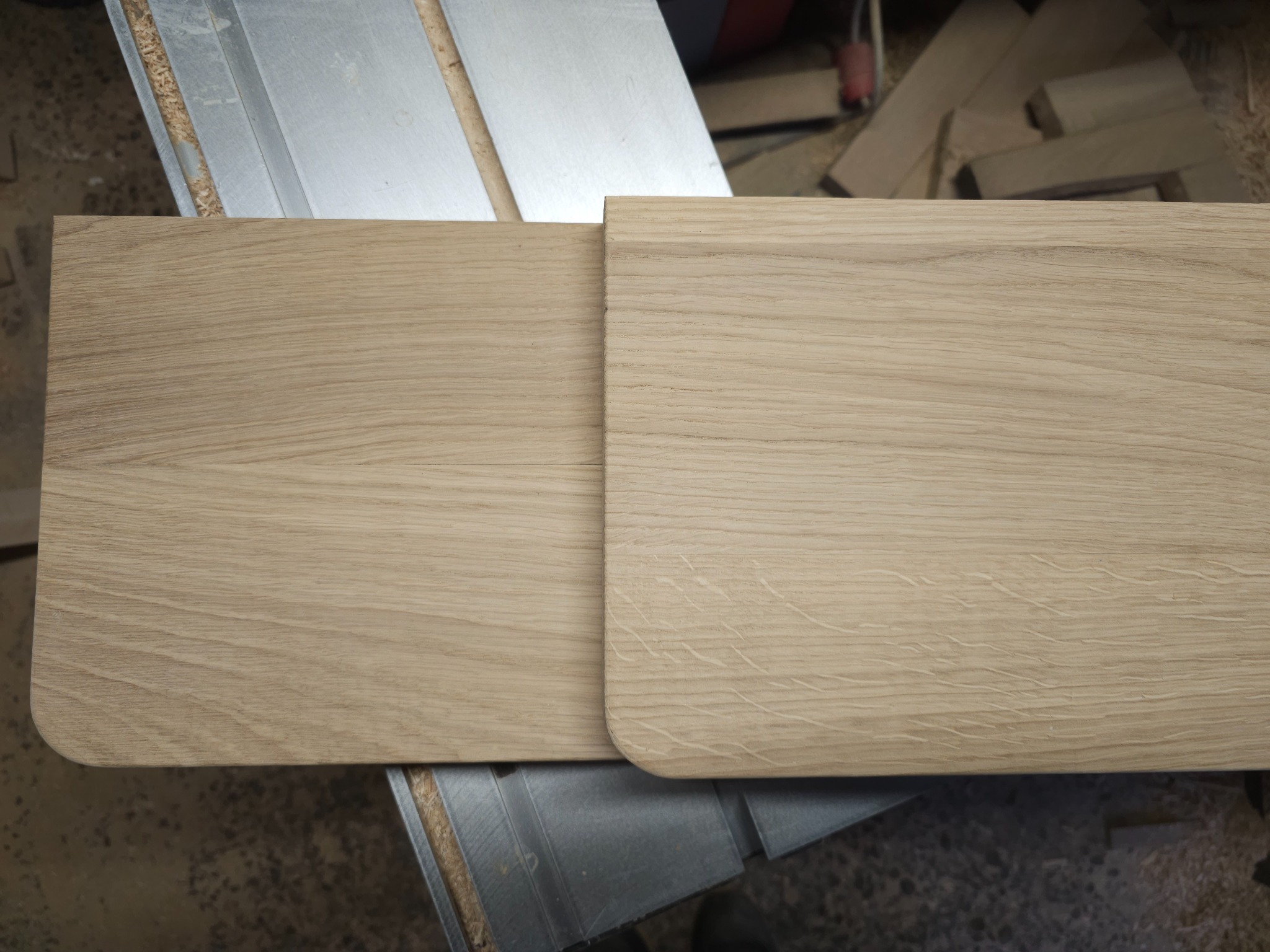 Dwa jasne, dębowe elementy drewniane z zaokrąglonymi krawędziami, leżące na metalowym blacie roboczym. Widoczne słoje drewna i drobne wióry.