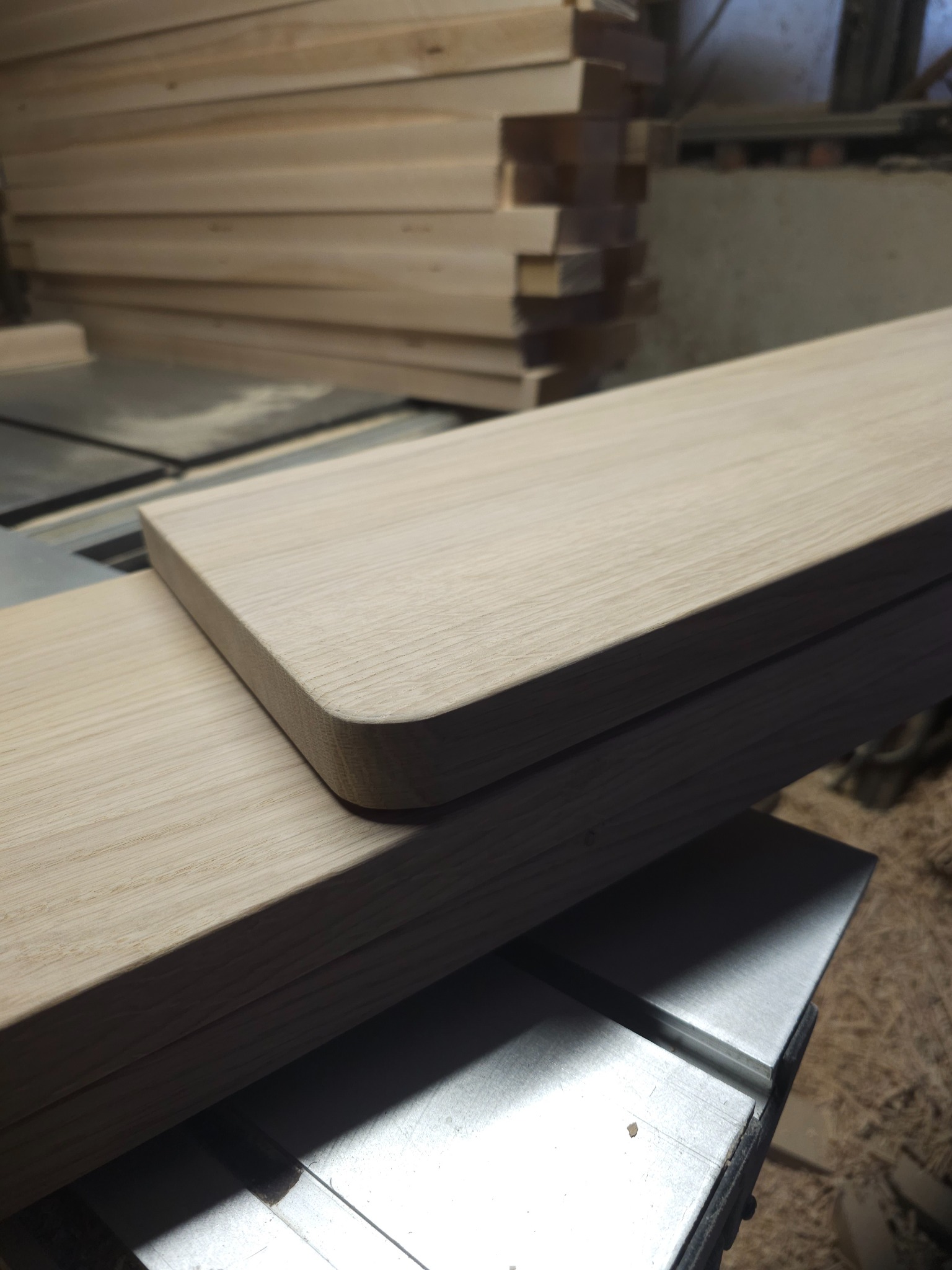 Obróbka drewna: Frezowana deska z zaokrąglonym rogiem na stole warsztatowym, w tle stos desek. Widoczne ślady obróbki.