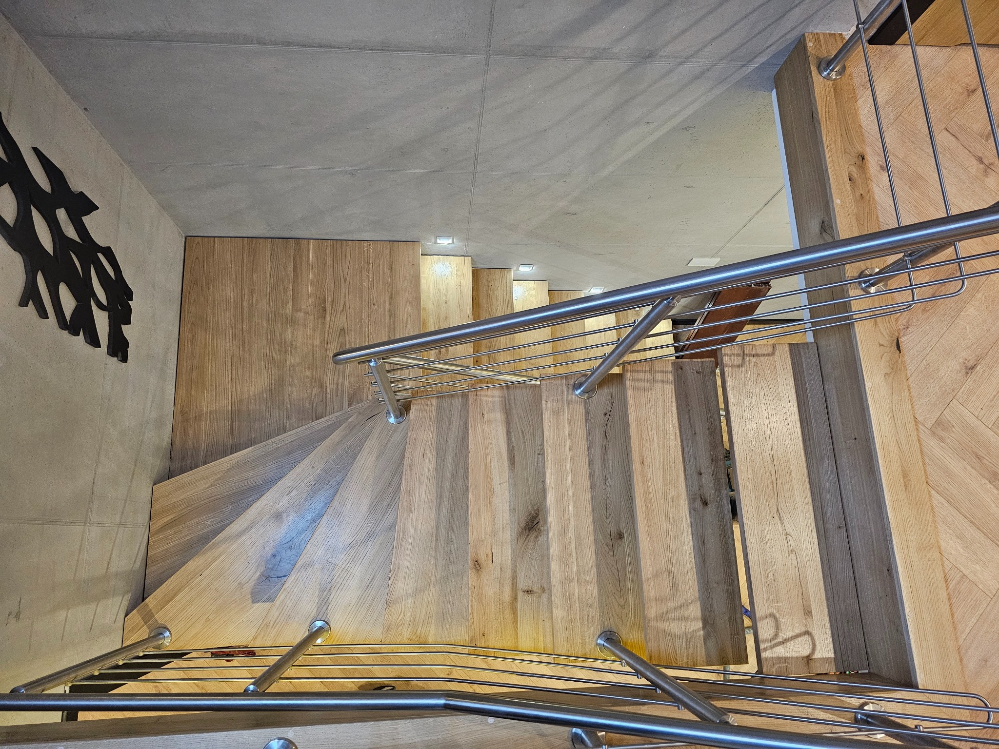 Spiralne schody z jasnego drewna z metalową balustradą, widok z góry, oświetlenie punktowe, dekoracja ścienna w kształcie drzewa, nowoczesny design.