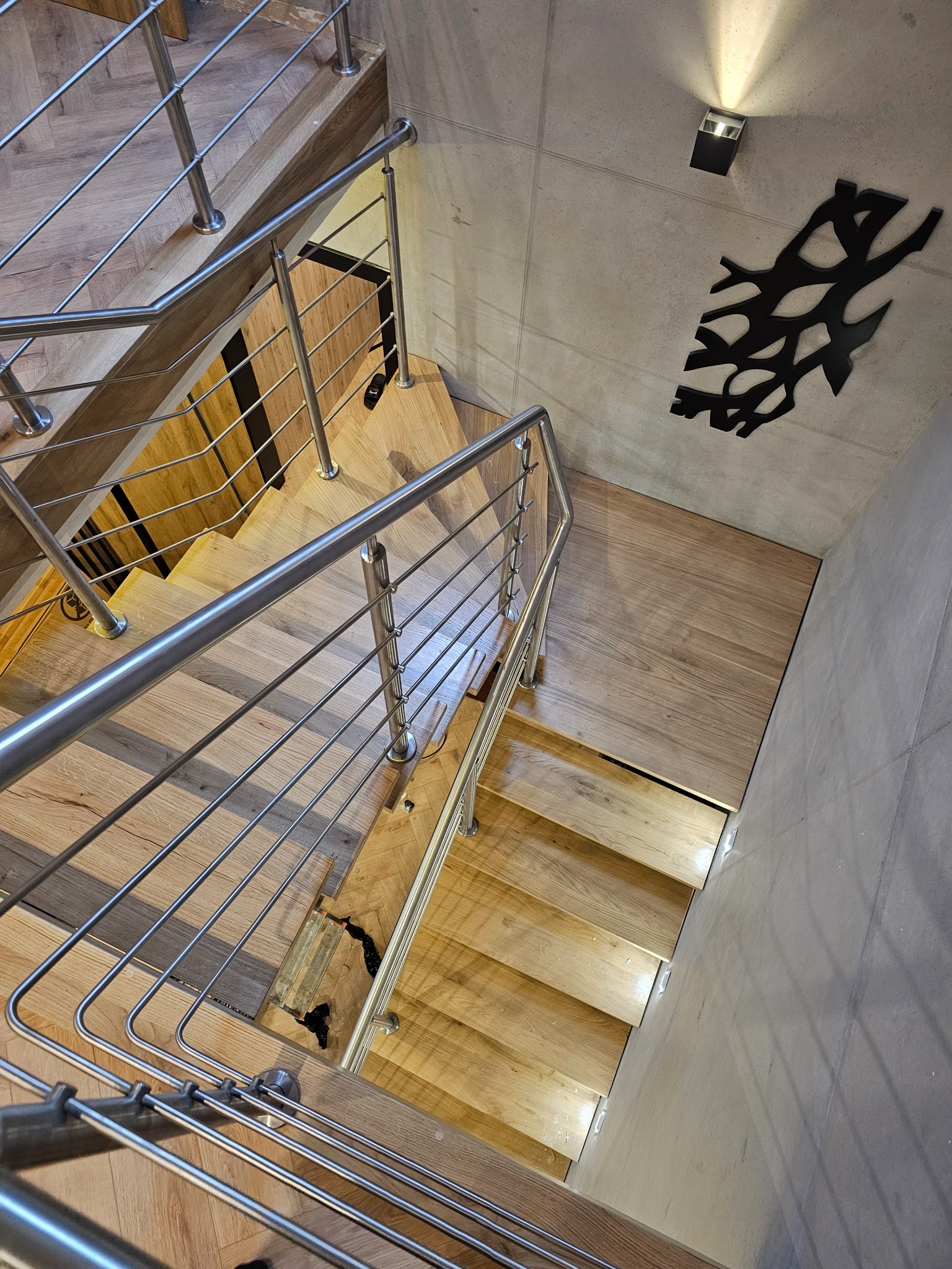 Nowoczesne schody z jasnego drewna z metalową balustradą, widok z góry. Na ścianie dekoracja w kształcie gałęzi i oświetlenie punktowe.