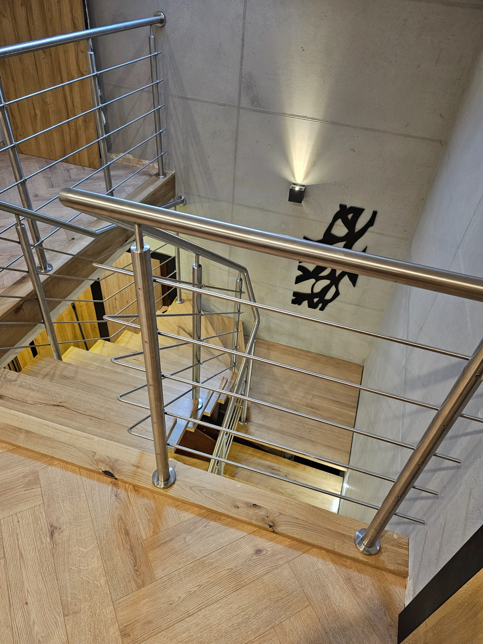 Nowoczesne schody z jasnego drewna ułożonego w jodełkę, z metalową balustradą. Widok z góry na dół, z oświetleniem punktowym na betonowej ścianie.
