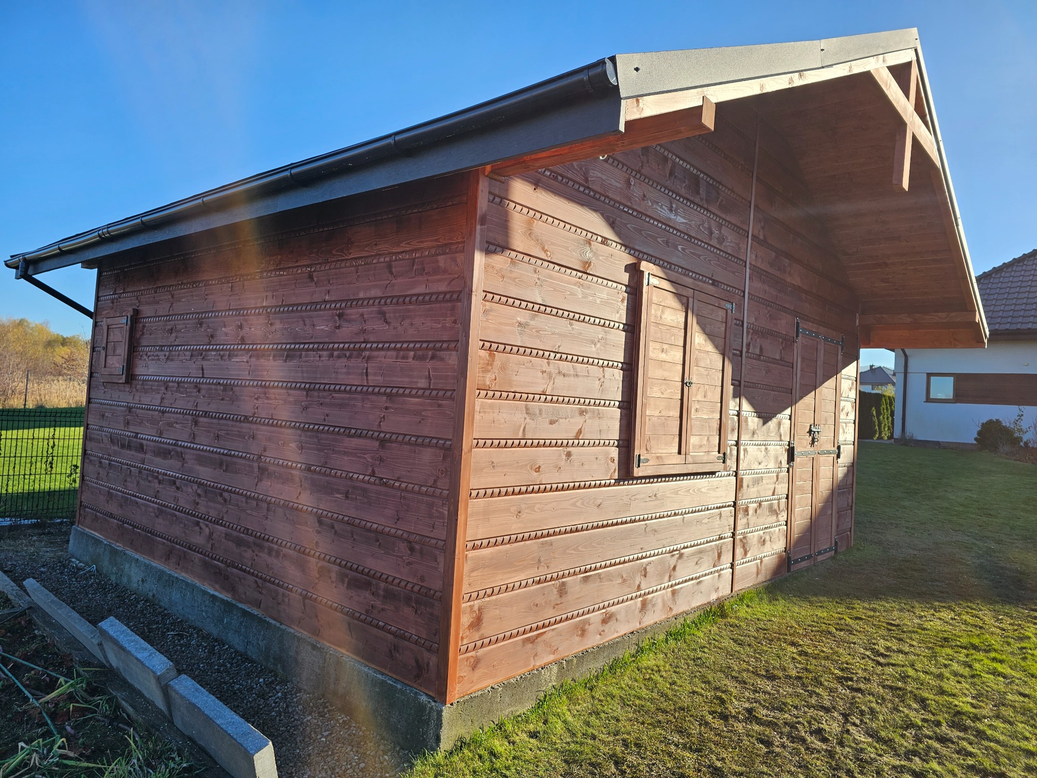 Drewniana altana z zamkniętymi okiennicami, wykończona w ciepłym brązie, na tle zielonego trawnika i błękitnego nieba. Widoczne detale zdobień na desce elewacyjnej.