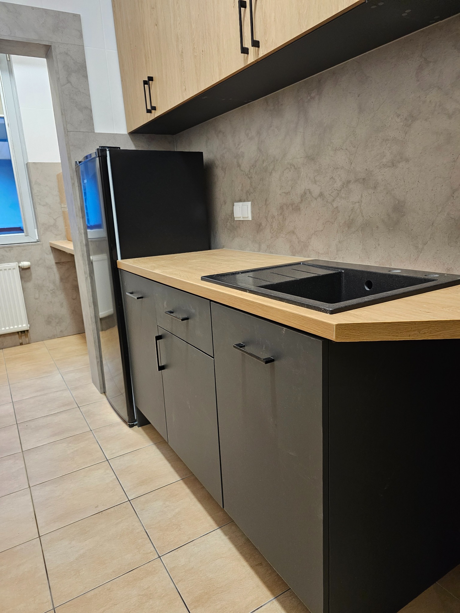 Nowoczesna kuchnia z czarnymi szafkami i drewnianym blatem, widok na zlew i lodówkę. Minimalistyczny design, szare ściany i jasne płytki podłogowe.