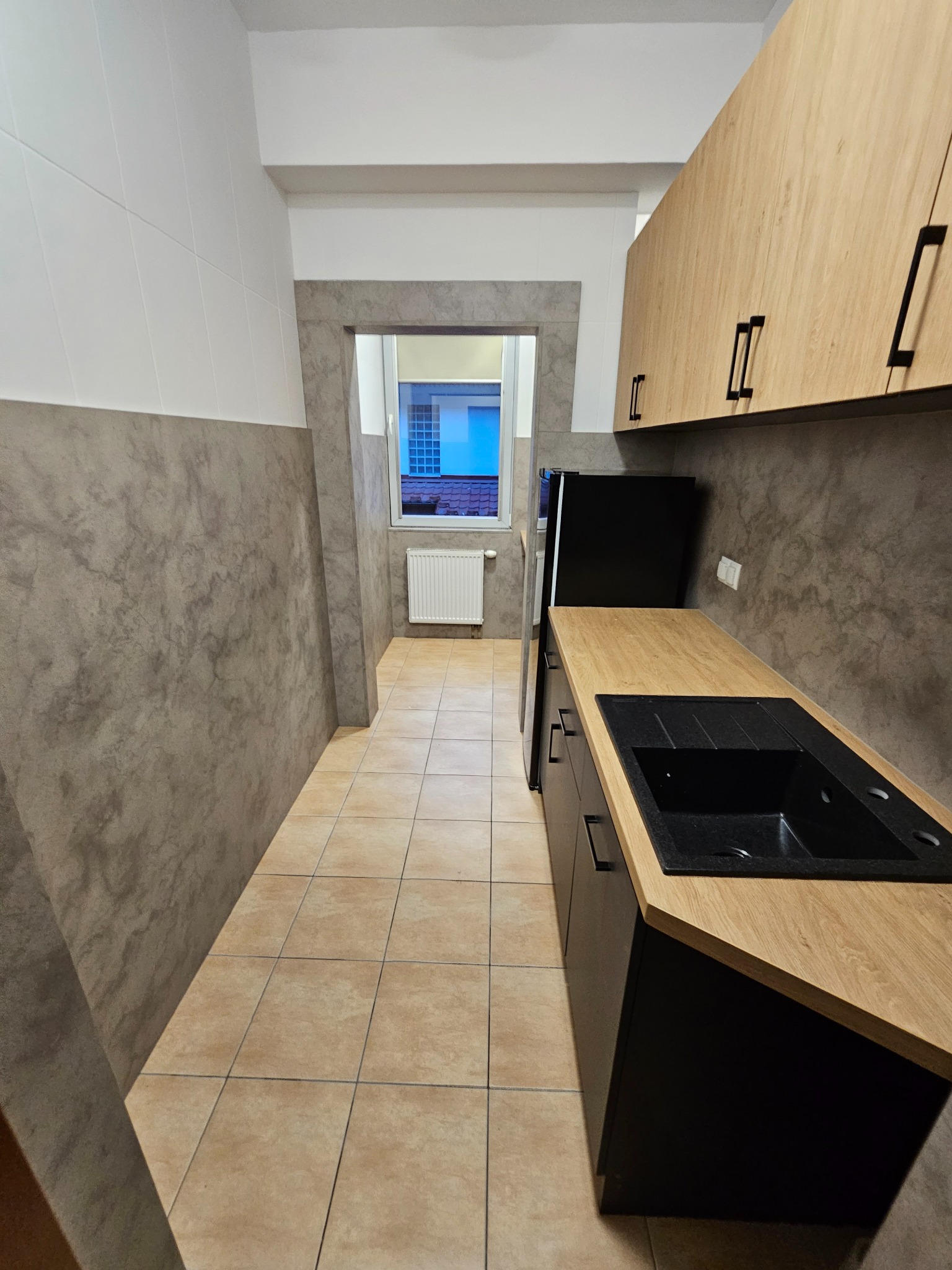Wąska kuchnia z zabudową: czarne szafki dolne z drewnianym blatem, jasne szafki górne. Widok na okno i grzejnik na końcu pomieszczenia. Szare ściany z efektem betonu.