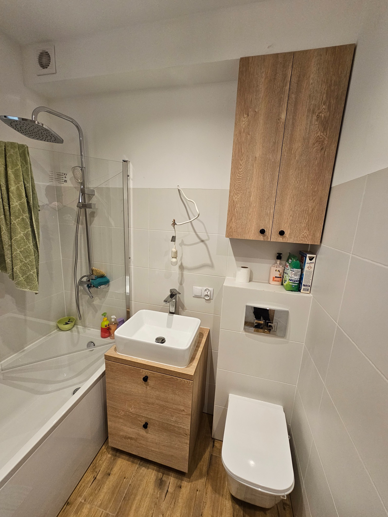 Nowoczesna łazienka z drewnianą szafką pod umywalką i wiszącą szafką. Biała armatura i płytki. Uwagę zwraca drewniany akcent w postaci szafek.