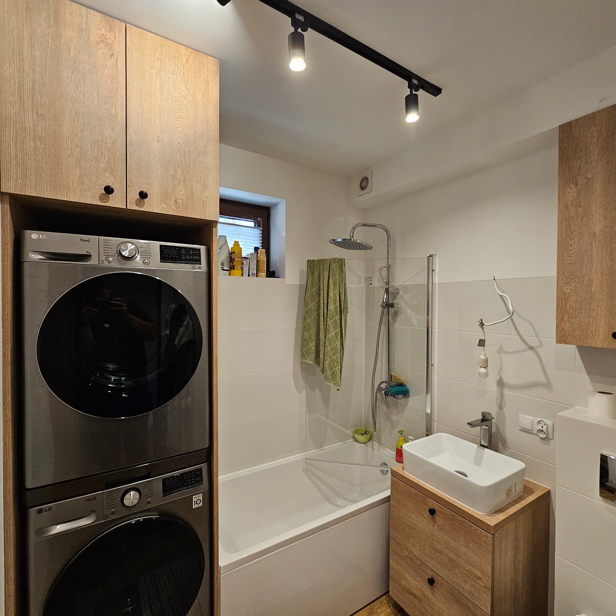 Funkcjonalna łazienka z pralką i suszarką w zabudowie z drewnianymi szafkami, białą wanną i umywalką nablatową. Oświetlenie punktowe na suficie.