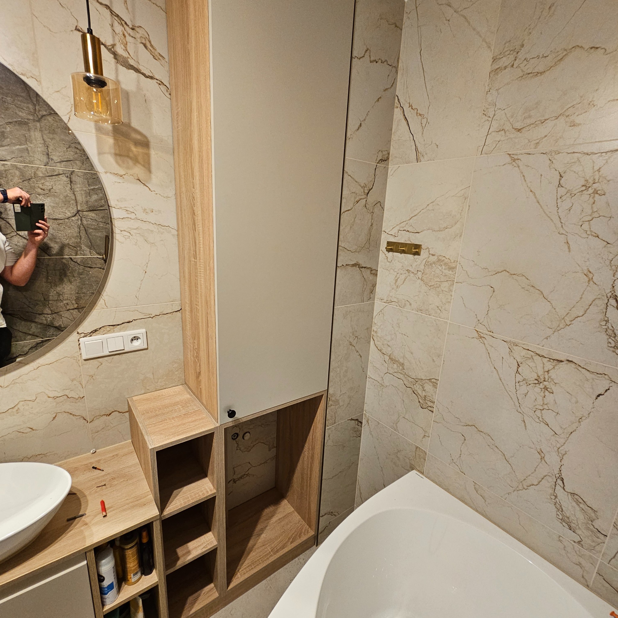 Nowoczesna łazienka z szafką na wymiar w odcieniach beżu i bieli, marmurowe płytki, okrągłe lustro z oświetleniem. Widoczna wanna i umywalka.