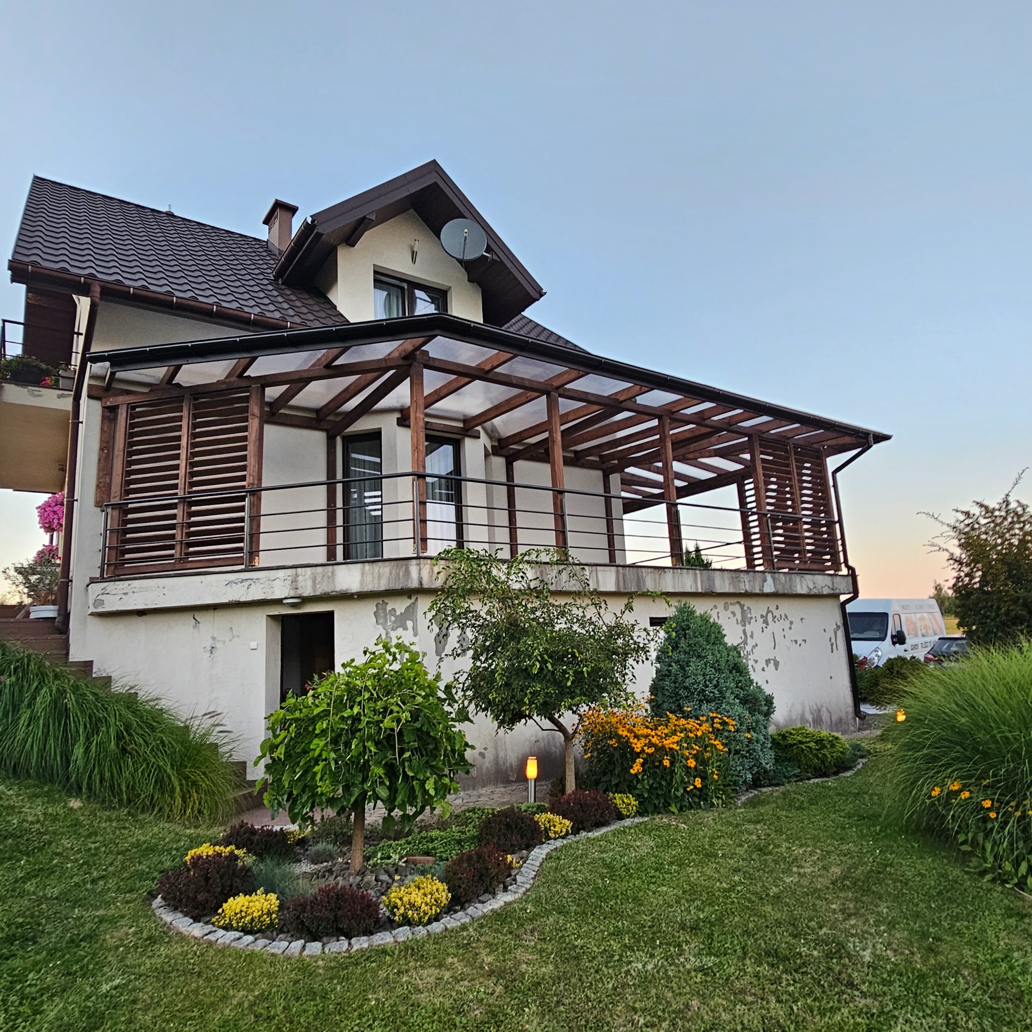 Dom z zadaszonym tarasem z drewnianą konstrukcją i balustradą. Widoczne elementy ogrodu i oświetlenie. Ujęcie z dołu, w tle błękitne niebo.