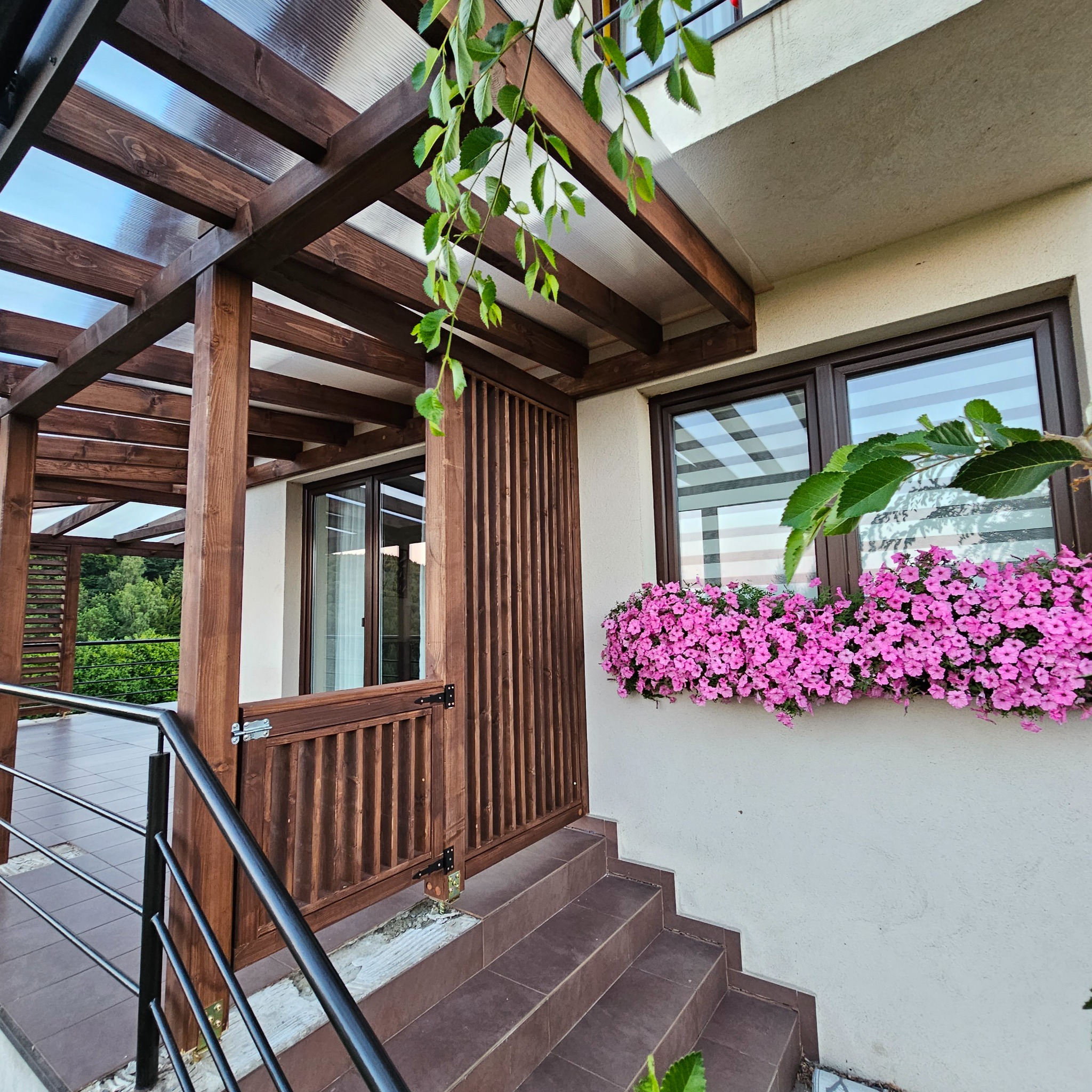 Zadaszona pergola z drewna i schody z płytek w Nowym Sączu. Metalowa balustrada, kwiaty w oknie. Widok z dołu, jasne światło dzienne.