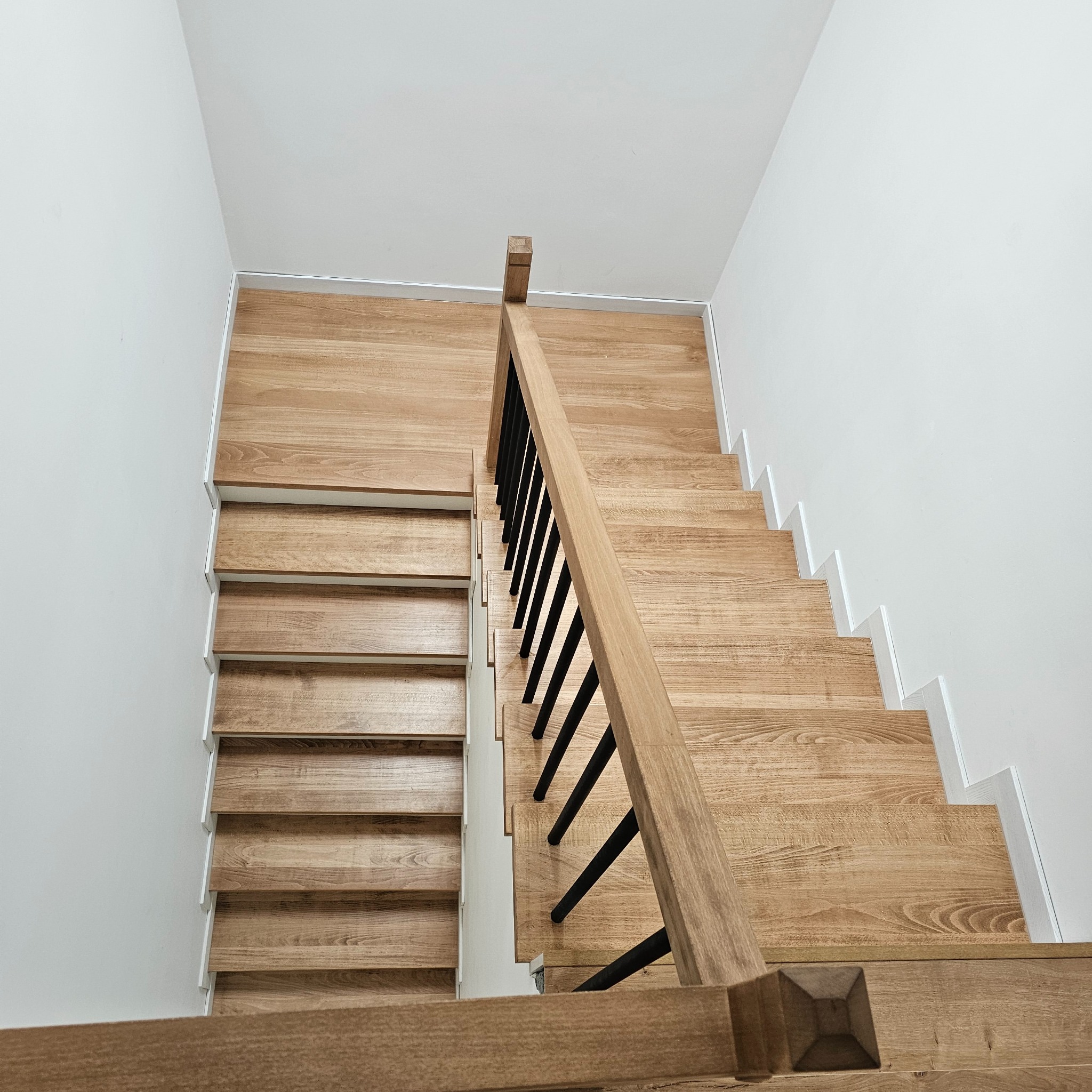 Nowoczesne schody z jasnego drewna z czarnymi balustradami, widok z góry, minimalistyczny design, białe ściany, proste wykończenie.