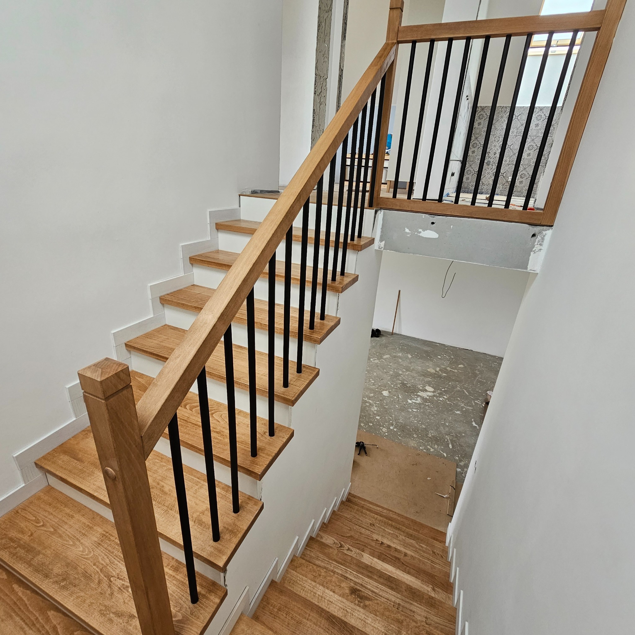 Drewniane schody z czarną balustradą w jasnym wnętrzu. Widok z góry na stopnie z wyraźnym usłojeniem drewna i białe podstopnice. Częściowo wykończone pomieszczenie w tle.