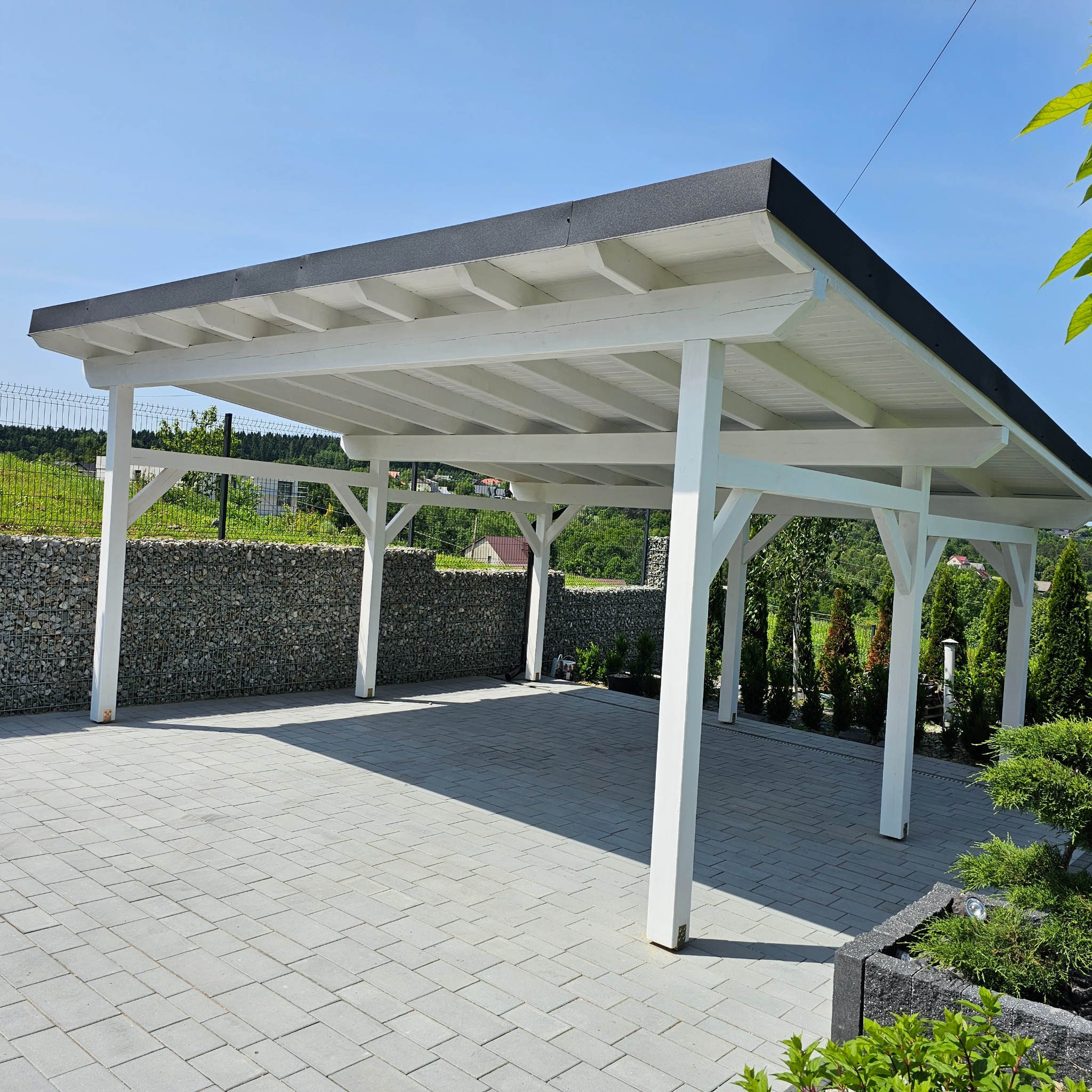 Biała, drewniana wiata garażowa z płaskim dachem na posesji z kostki brukowej. W tle ogrodzenie z kamieni i zieleń. Słoneczny dzień, błękitne niebo.