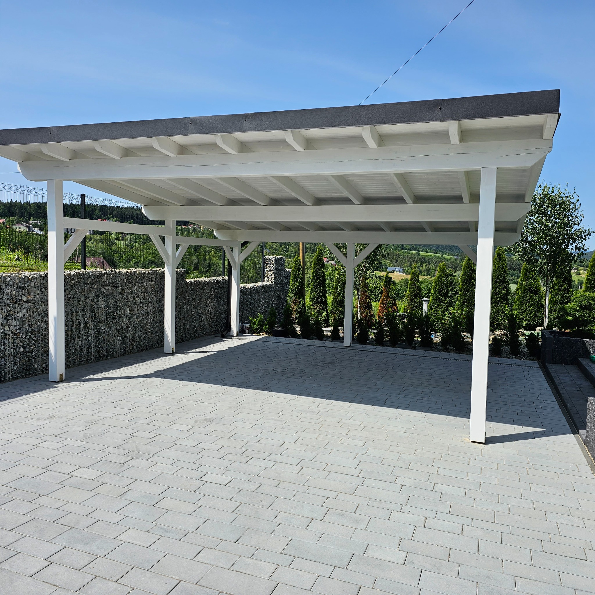 Nowoczesna, biała wiata garażowa z płaskim dachem na posesji z kostki brukowej. W tle zielony krajobraz i ogrodzenie z gabionów.