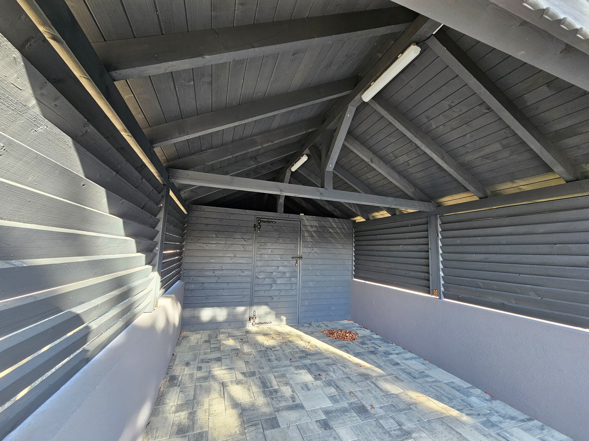 Szara drewniana konstrukcja altany z zamykanymi drzwiami w Nowym Sączu, z widocznym oświetleniem i fragmentem kostki brukowej na podłodze.