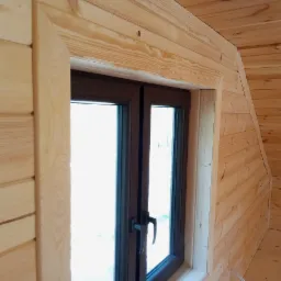 Wnętrze domu wykończone drewnem, z brązowym oknem z podwójnymi szybami i drewnianą ramą okienną, widoczne łączenie paneli ściennych pod kątem.