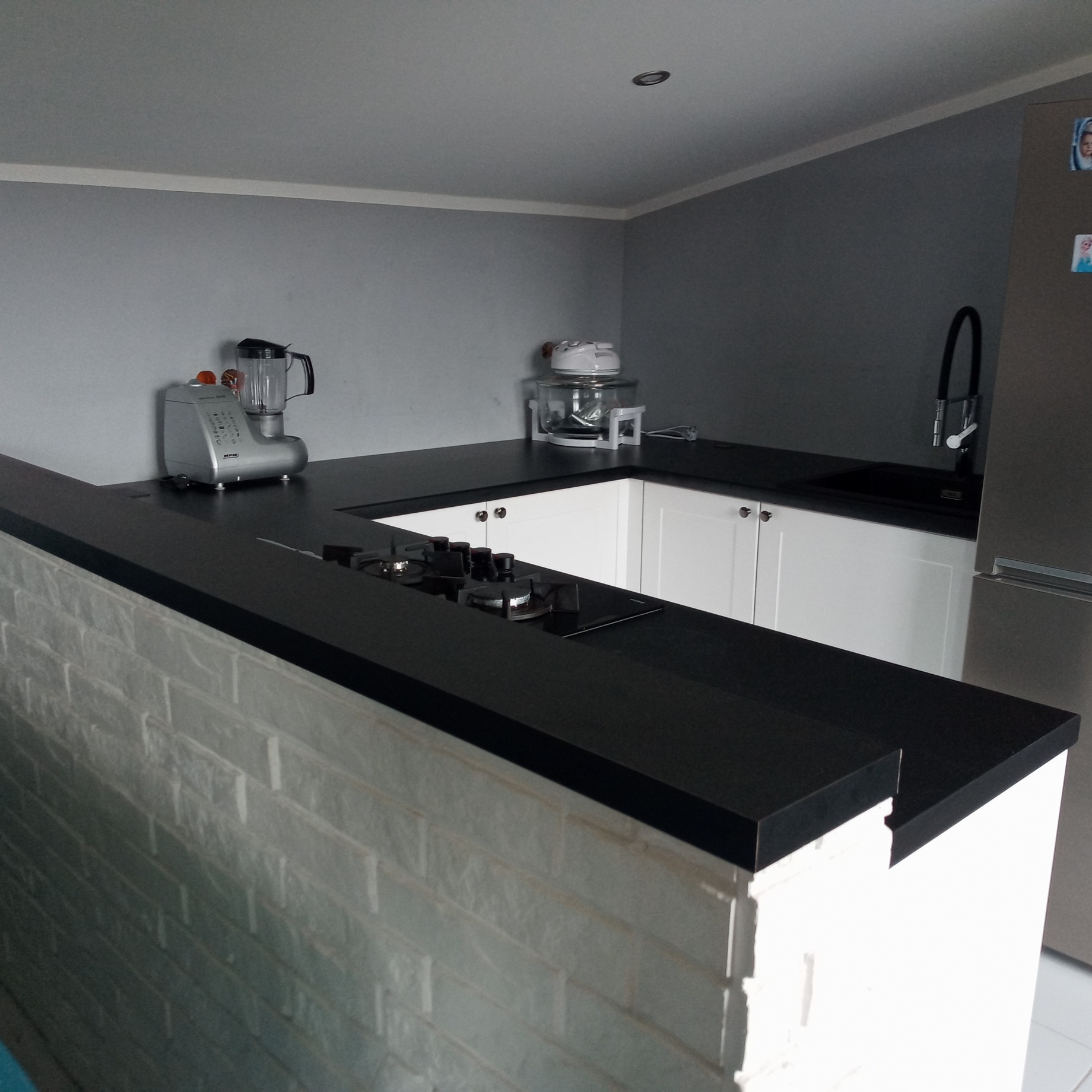 Nowoczesna kuchnia z czarnym blatem, białą zabudową i ścianą z białej cegły dekoracyjnej, widoczny sprzęt AGD i płyta grzewcza.