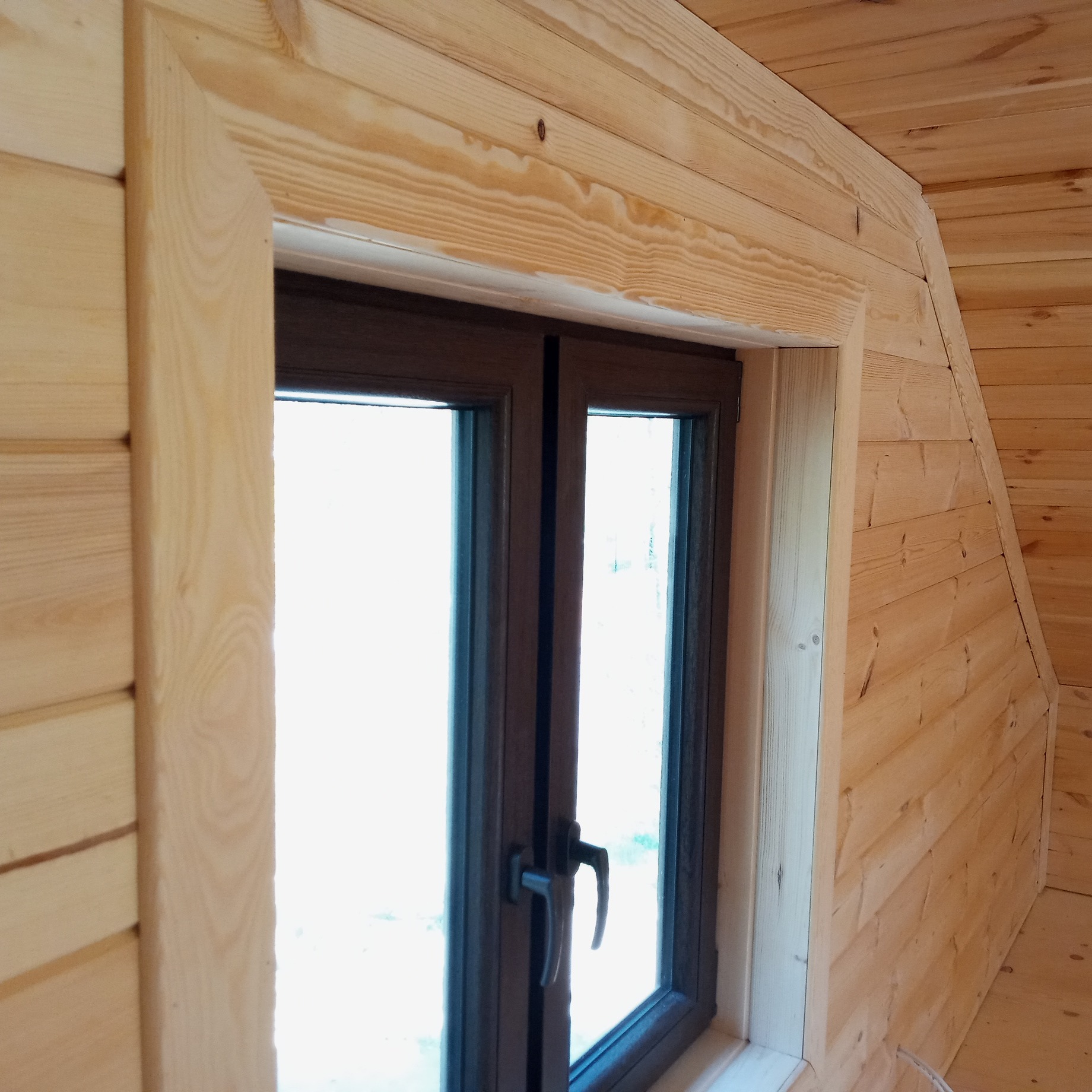 Wnętrze domu wykończone drewnem, z brązowym oknem z podwójnymi szybami i drewnianą ramą okienną, widoczne łączenie paneli ściennych pod kątem.