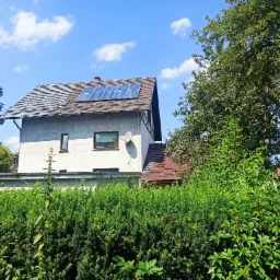 Dom jednorodzinny z panelami słonecznymi na dachu, widoczny zza żywopłotu w słoneczny dzień.