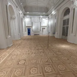 Rozległa sala z nowo ułożoną, wzorzystą parkietową podłogą w odcieniach beżu i brązu, wysokie białe ściany z dekoracyjnymi sztukateriami i łukowymi oknami, widok perspektywiczny.