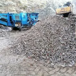 Kruszywo skalne z niebieską kruszarką i żółtą koparką gąsienicową w tle, obok fragmentu ceglanej ściany.