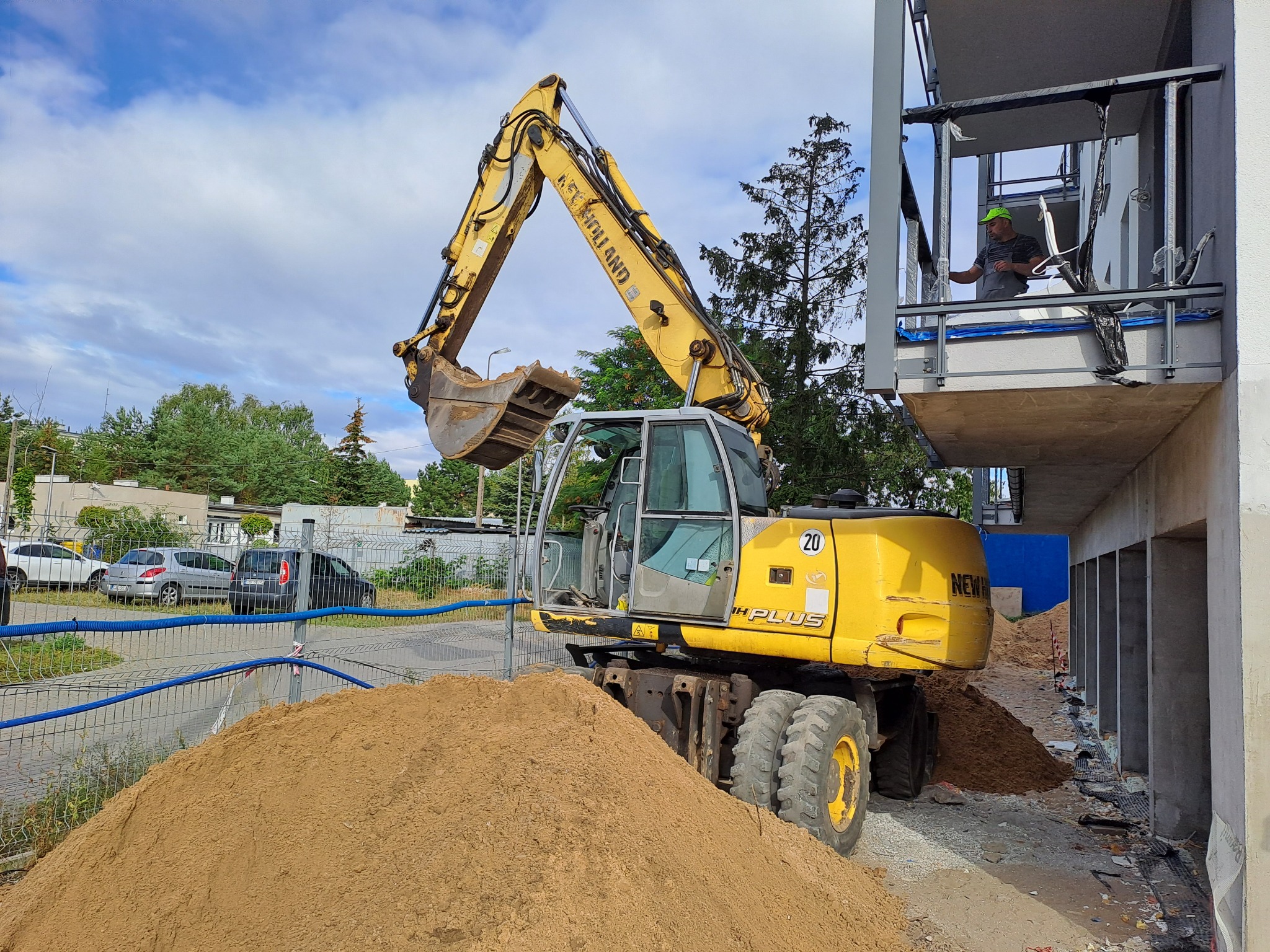 Żółta koparka kołowa na placu budowy z hałdą piasku. W tle budynek w stanie surowym i pracownik na balkonie. Błękitne niebo z chmurami.