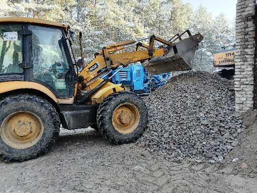 Żółta ładowarka JCB z pełną łyżką żwiru obok pryzmy tego samego materiału; w tle niebieska kruszarka i fragment ceglanego budynku.