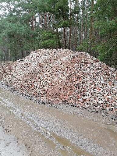 Góra czerwono-szarego gruzu lub tłucznia leżąca obok błotnistej drogi gruntowej, w tle widoczny las sosnowy.