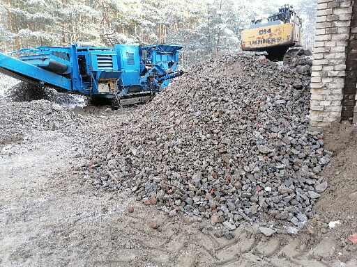 Kruszywo skalne z niebieską kruszarką i żółtą koparką gąsienicową w tle, obok fragmentu ceglanej ściany.