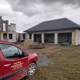 Usługi Ciesielsko-Budowlane DAR-BUD Dariusz Czerwiński - Budowa domu w stanie surowym otwartym z widocznym dachem i ścianami z bloczków. Na pierwszym planie czerwony samochód z logo firmy budowlanej.