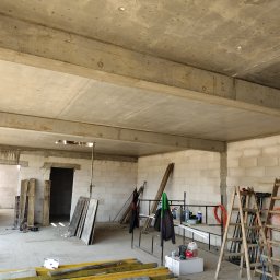 Usługi Ciesielsko-Budowlane DAR-BUD Dariusz Czerwiński - Surowy stan domu w budowie: ściany z betonu komórkowego, betonowy strop z widocznymi belkami, rusztowania i drabiny wewnątrz. Widoczne narzędzia i materiały budowlane.