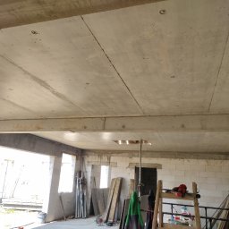 Usługi Ciesielsko-Budowlane DAR-BUD Dariusz Czerwiński - Surowy betonowy strop w nowym budynku z pustaków, widoczne otwory i belki nośne, przygotowany do dalszych prac wykończeniowych. W tle okna i materiały budowlane.