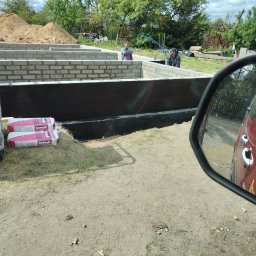 Usługi Ciesielsko-Budowlane DAR-BUD Dariusz Czerwiński - Budowa fundamentów: ściany z bloczków, izolacja pionowa, worki z zaprawą Ceresit. Dwóch pracowników przy pracy, widok zza lusterka samochodu.