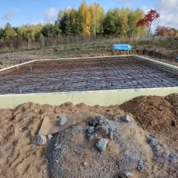 Przygotowany zbrojony fundament pod budowę domu, z widoczną siatką zbrojeniową i izolacją termiczną, otoczony ziemią i zaroślami w tle.