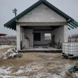 Surowy stan budynku gospodarczego z białych bloczków, z widocznym szkieletem dachu, stertą gruzu przed wejściem i zbiornikiem na wodę z boku na placu budowy pokrytym resztkami śniegu.