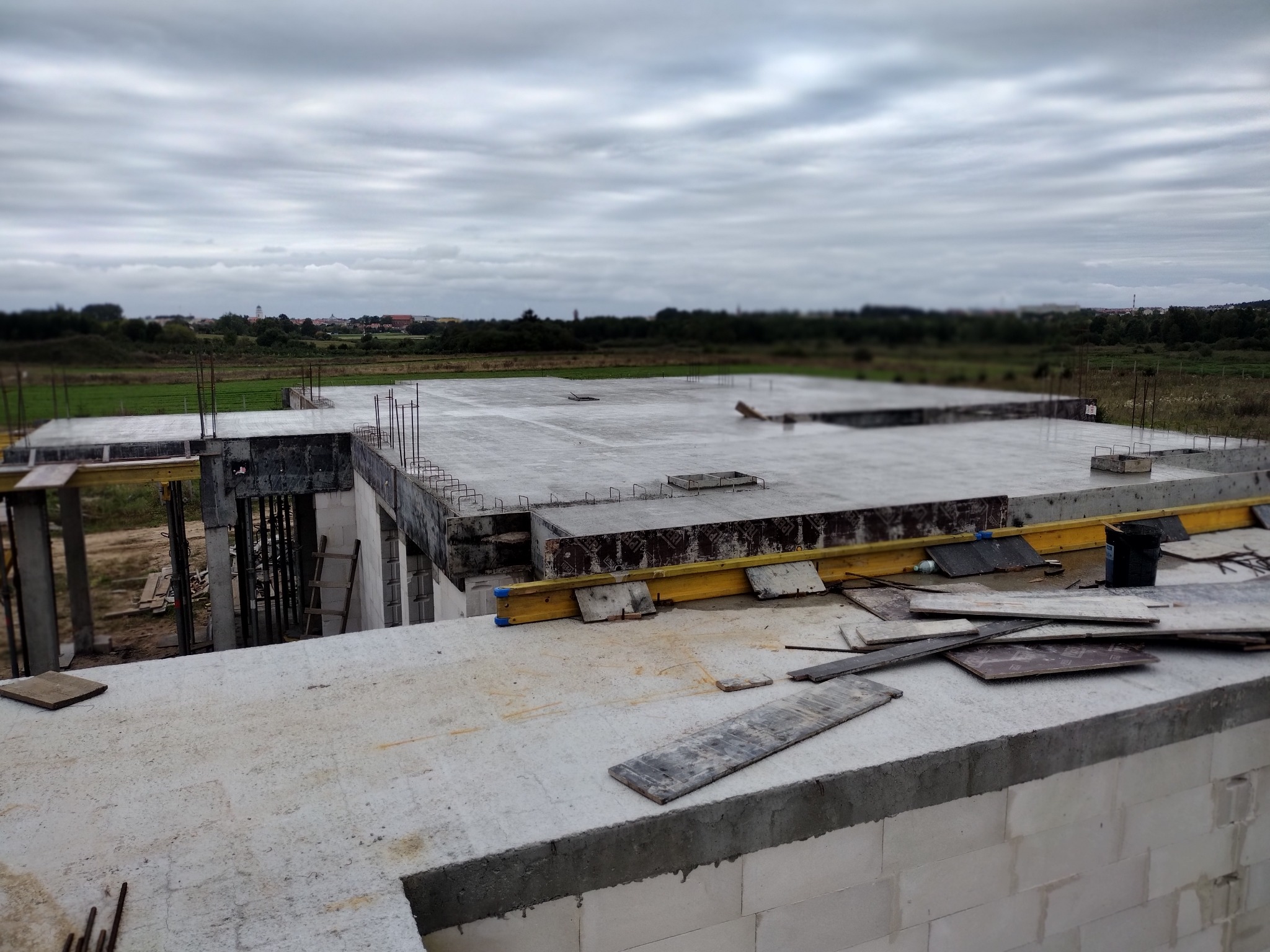 Wysoki kąt na betonowy fundament domu w trakcie budowy, z widocznym zbrojeniem i deskowaniem. W tle zieleń i pochmurne niebo.