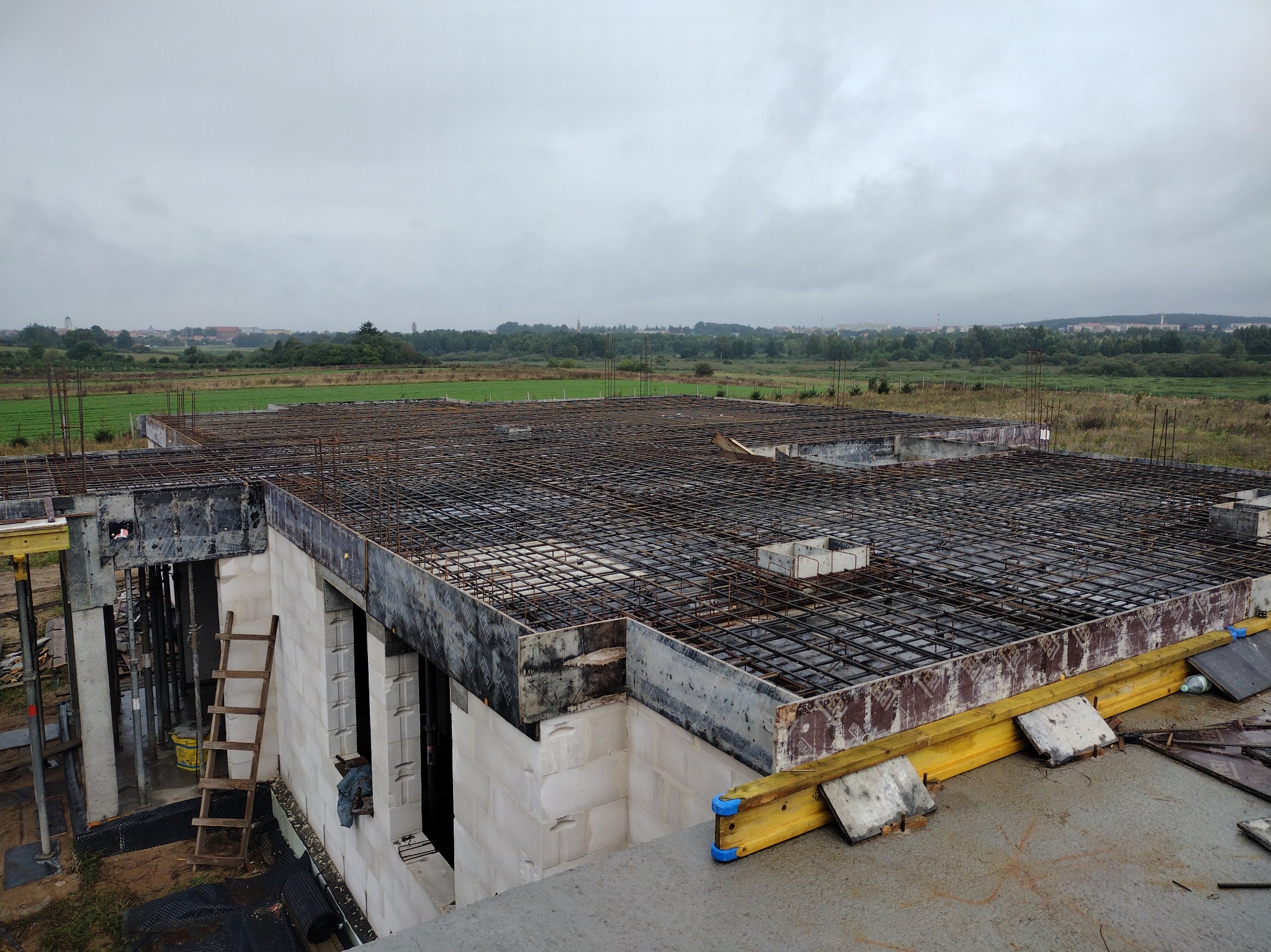 Budowa domu z betonu komórkowego: zbrojenie stropu z widoczną drabiną i deskowaniem, w tle krajobraz pod pochmurnym niebem. Etap przed zalaniem betonem.