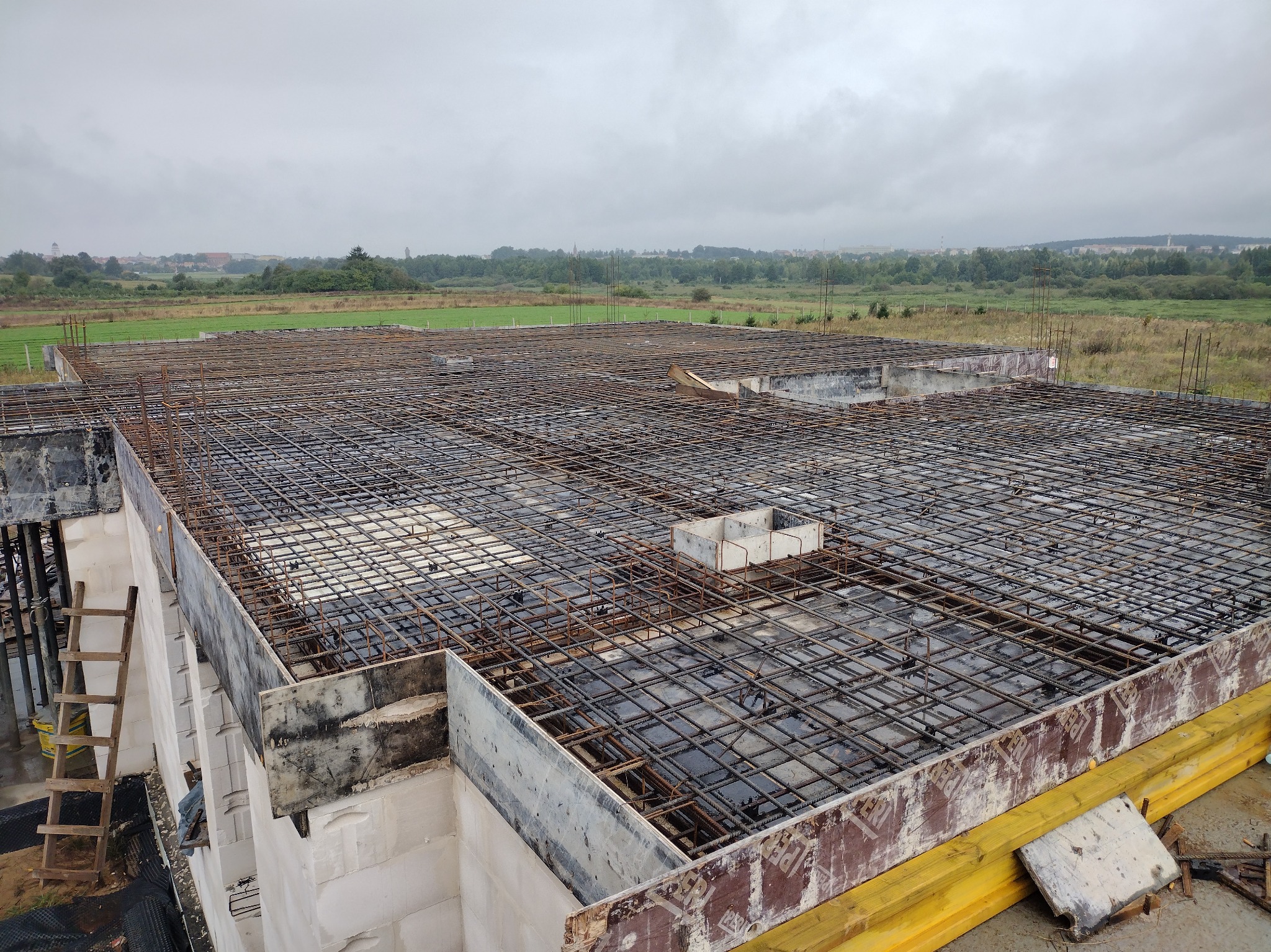 Zbrojenie stropu domu jednorodzinnego z widoczną konstrukcją szalunkową i drewnianą drabiną. W tle rozległy krajobraz z zielenią i zabudowaniami.